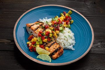Delicious grilled salmon steak on a blue plate with jasmine rice and sauce made of mango, bell pepper, onion and avocado. Top notch food on a wooden table.
