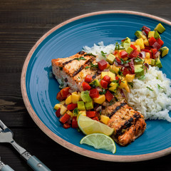 Grilled fillet of salmon with rice garnish and exotic fruit sauce made of mango, avocado, pepper and onion on a rustic wooden table.