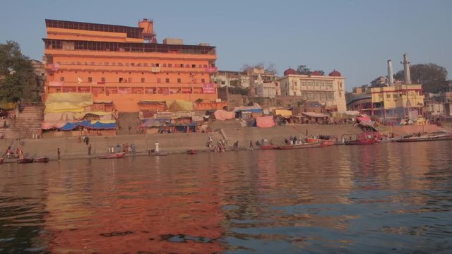 View of buildings of temples with ancient architecture and ghats of Varanasi, holy Hindu town, as seen from a moving boat pov in the river Ganges at sunrise with oar