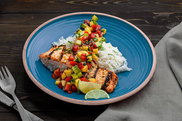 Grilled salmon fillet with fruit sauce made of mango, bell pepper, onion and avocado with basmati rice garnish on a blue plate. Delicious seafood meal on a table.