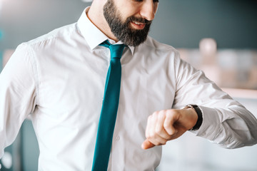 Close up of attractive bearded businessman in shirt and tie looking at wristwatch while standing in office. It's time to inspire.