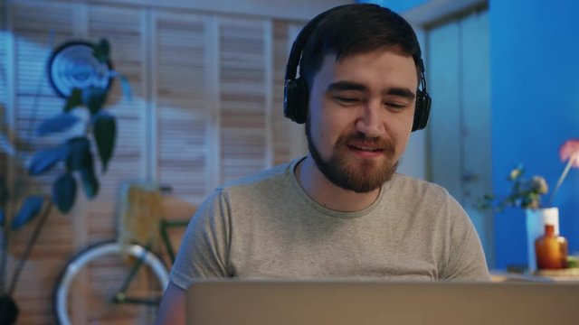 Cute young man with beard sitting at laptop, talking to foreign friends. Language exchange site. Online. Indoors. Conversation. Home. Night. Different time zones around world.