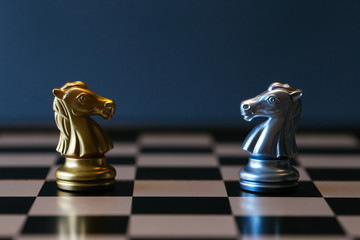 Closeup golden and silver horse knights places against each other on chessboard against gray...