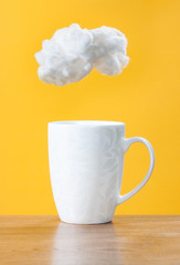 White cup on a wooden table. White cloud of cotton wool over a cup on a yellow background. Steam raising concept