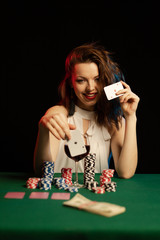 Emotional young lady in a white blouse drinking wine from a glass and playing cards on a table on...