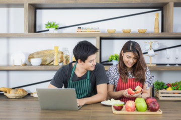 Happy young Asian couple sharing relax times in kitchen room, husband turn and talk to wife who preparing apple for light snack at afternoon tea time