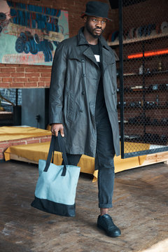 Fashionable young man in black waxed trench coat, bucket hat and with blue bag, walking in loft interior room with painting and chain link.