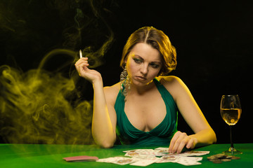 A young lady in a green dress smokes a cigarette and blows smoke and plays cards on a table on...
