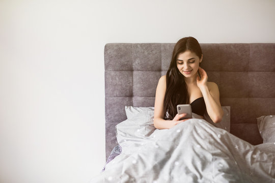 Smiling young woman in sexy lingerie holding mobile phone while lying on bed at home