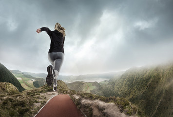 Higher than clouds and faster than wind. Young woman in fitness wear and shoes is running and jumping near mountains, meadows and lake in cloudly day. Sport, lifestyle and healthy life concept.