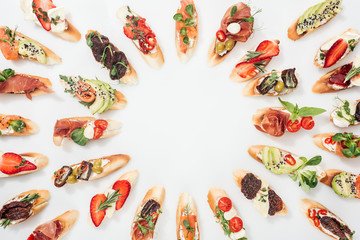 Fototapeta na wymiar top view of round frame made of italian bruschetta with salmon, prosciutto, herbs and various fruits with vegetables on white