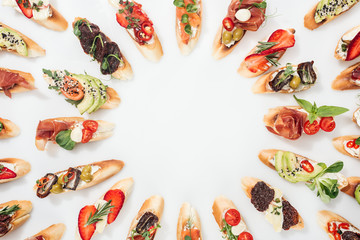 top view of round frame made of italian bruschetta with salmon, prosciutto, herbs and various fruits with vegetables on white
