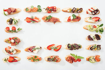 top view of frame made of traditional italian bruschetta with prosciutto, salmon, fruits, vegetables and herbs on white