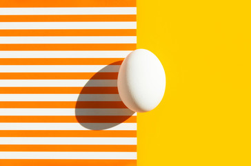 One white egg on duotone yellow orange and white striped background. Easter concept. Hard light harsh shadow. Trendy minimalist pop art style. Bright vivid colors. Holiday card poster