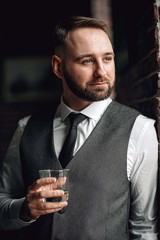successful young businessman in a vest and tie. with a stylish and stylish haircut and beard. holding a glass of whiskey in his hands