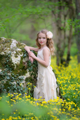 A cute young girl is in the forest on the edge of the forest with yellow flowers. She has blond curly hair, she's wearing a beautiful dress.