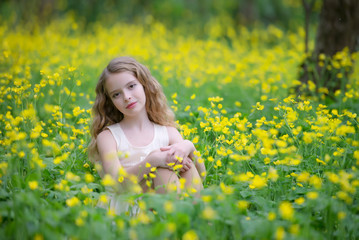 Naklejka na ściany i meble A cute young girl is in the forest on the edge of the forest with yellow flowers. She has blond curly hair, she's wearing a beautiful dress.