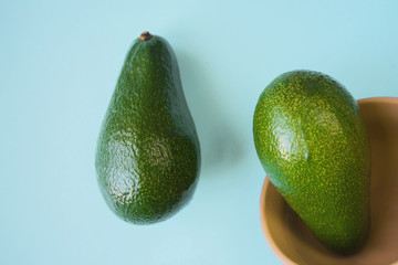 one ripe green avocado lies on a blue background, the second in a deep beige plate beside