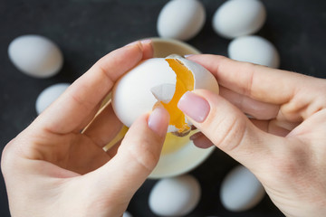 Fototapeta na wymiar female hands open the shells of a fresh white chicken egg on a dark background with other eggs and yolk and white are visible