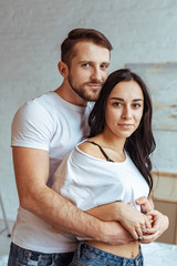 handsome man hugging beautiful and brunette woman in t-shirt and looking at camera