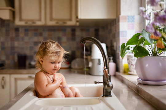 Baby Taking Bath In Kitchen Sink. Child Playing With Water And Soap Bubbles In Sunny Kitchen With Window. Water Fun For Kids. Hygiene And Skin Care For Children. Kitchen And Bath Room Interior