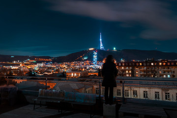 Fototapeta na wymiar Georgia, Tbilisi - 05.02.2019. - Night cityscape view with human silhouette standing on the rooftop. Famous landmarks illuminated - Image