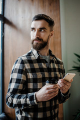 Bearded attractive man, handsome hipster bearded guy holding mobile cell phone and cup of morning coffee in new modern coffee shop via the searching network. Free wi-fi and technologies concept