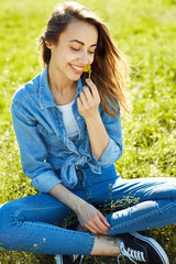portrait of attractive woman in jeans sitting in a park on the grass at sunny summer day. joyful smiling woman holds a dandelion flower in hand and sniffs it