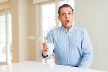 Middle age man drinking coffee in the morning at home scared in shock with a surprise face, afraid...