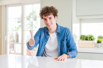 Young handsome man wearing casual denim jacket at home smiling friendly offering handshake as greeting and welcoming. Successful business.