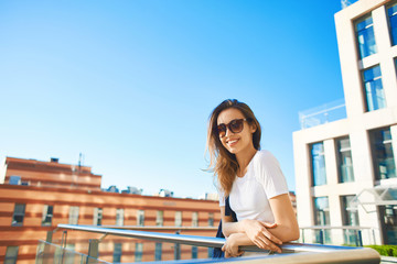 portrait of a young smiling attractive woman in white t-shirt with small city backpack at sunny day on city building and blue sky background. woman poses in cityscape.