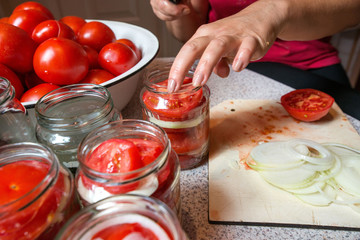 Canning fresh tomatoes with onions in jelly marinade. Woman hands putting red ripe tomato slices and onion rings in jars. Basil, parsley leaves on top of onions. Vegetable salads for winter