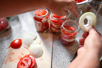 Canning fresh tomatoes with onions in jelly marinade. Woman hands putting red ripe tomato slices and onion rings in jars. Basil, parsley leaves on top of onions. Vegetable salads for winter