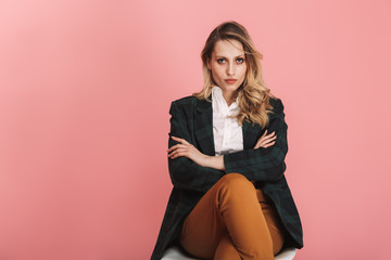 Photo of serious businesswoman sitting in chair and looking on camera isolated over pink background