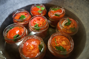 Canning fresh tomatoes with onions in jelly marinade. Conservation and cooking. Jars boiling in water to kill bacteria. Sealing process.  Basil, parsley leaves on top. Vegetable salads for winter
