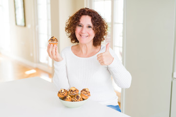 Senior woman eating chocolate chips muffins happy with big smile doing ok sign, thumb up with fingers, excellent sign