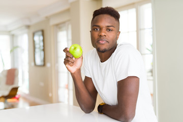 Young african american man eating fresh green apple with a confident expression on smart face thinking serious