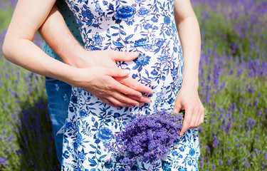 Cropped image of pregnant woman in blue flowery dress and her husband hugging the tummy in lavender field