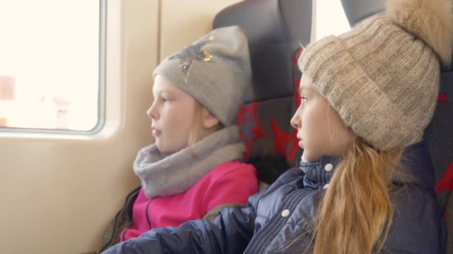 Two teen girls in winter clothes jackets and caps are travelling by train and looking at window. Side view.