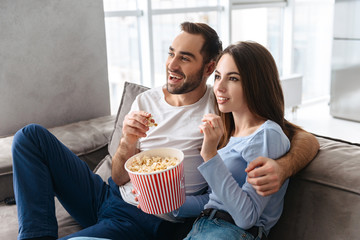 Image of adorable couple eating popcorn from bucket while sitting on couch indoor and watching movie