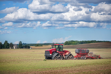 Fototapeta premium Agricultural background with red tractor pulling plow, throwing dust in air. Combine harvester at wheat field. Heavy machinery during cultivation, working on fields. Dramatic sky, rain, storm clouds 