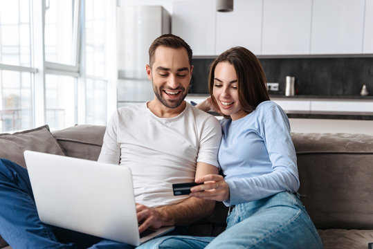 Image of lovely couple using laptop and credit card together for online shopping while sitting on sofa at home