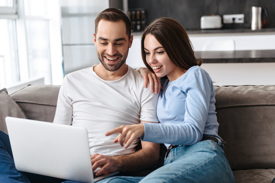 Image of happy couple using laptop together while sitting on sofa in living room at home