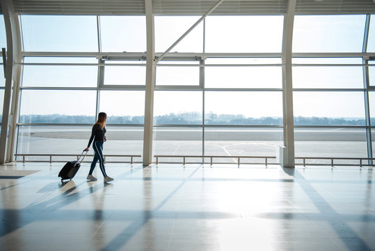 Young female pasenger in casual clothes with luggage walking near big windows in airport terminal hall. Waiting for a flight. Walking at the gate