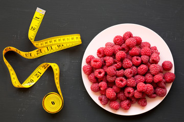 Raw raspberries on plate, measuring tape on black background, top view. Healthy eating and dieting....
