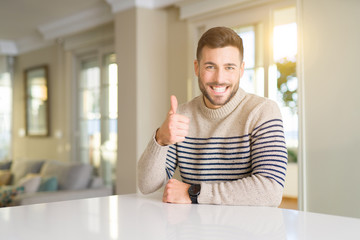 Young handsome man at home doing happy thumbs up gesture with hand. Approving expression looking at the camera showing success.