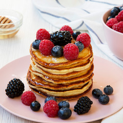 Homemade pancakes with berries and honey on a pink plate over white wooden surface, side view. Close-up.