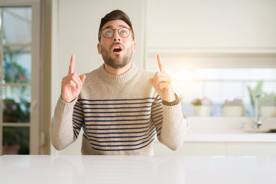 Young handsome man wearing glasses at home amazed and surprised looking up and pointing with fingers and raised arms.