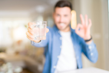 Young handsome man drinking a glass of water at home doing ok sign with fingers, excellent symbol