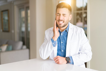 Young handsome doctor man at the clinic touching mouth with hand with painful expression because of toothache or dental illness on teeth. Dentist concept.